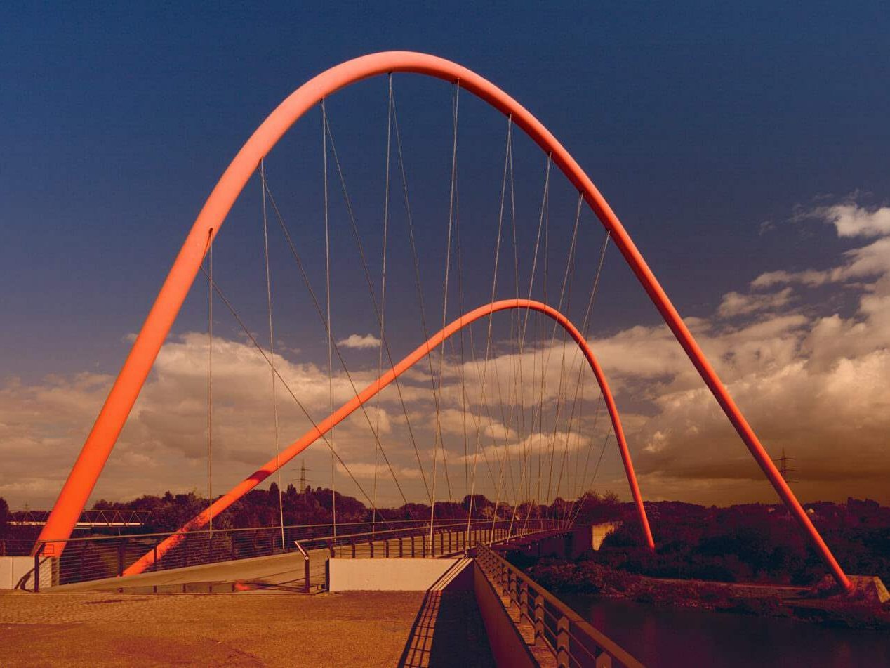 Eine rote, geschwungene Brücke ist zusehen, die über Bahnschienen verläuft.