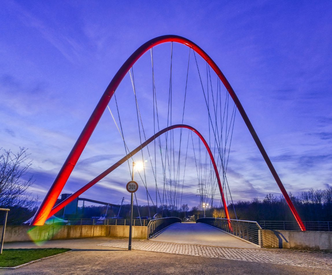 Eine rote, geschwungene Brücke ist zusehen, die über Bahnschienen verläuft.